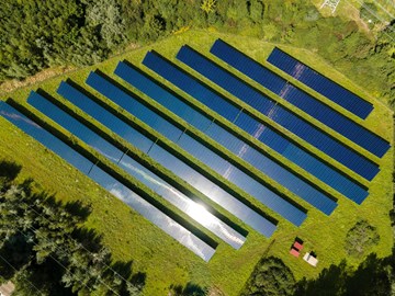 Solarpark Schkopau von oben Das Bild zeigt eine Luftaufnahme eines Solarparks mit mehreren Reihen von Solarmodulen, die auf einer grasbewachsenen Fläche installiert sind. Die Solarpaneele sind in parallelen Linien angeordnet. Auf einem der Module spiegelt sich das Sonnenlicht.