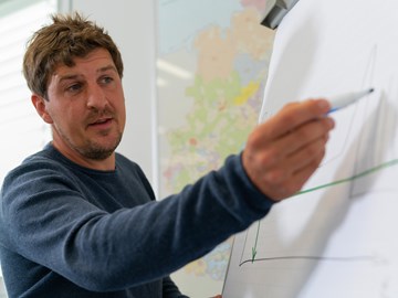 Mitarbeiter an Flipchart Eine Person zeigt mit einem Marker auf ein Diagramm auf einem Flipchart. Das Diagramm enthält Achsen und Linienverläufe, jedoch sind die genauen Inhalte nicht klar erkennbar. Im Hintergrund ist eine Karte zu sehen.