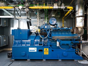 BHKW im Container Das Bild zeigt eine blaue Industriemaschine in einer Anlage. An der Maschine sind verschiedene Rohre, Ventile und elektrische Komponenten angeschlossen. Im Hintergrund befinden sich metallene Wände und weitere Ausrüstungsgegenstände.