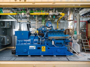 BHKW im Container Das Bild zeigt eine blaue Industriemaschine in einer Anlage. An der Maschine sind verschiedene Rohre, Ventile und elektrische Komponenten angeschlossen. Im Hintergrund befinden sich metallene Wände und weitere Ausrüstungsgegenstände.