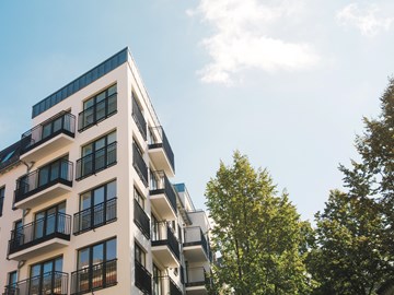 mehrstöckiges Wohngebäude Ein modernes, mehrstöckiges Wohngebäude mit hellen Fassaden und vielen Balkonen steht vor blauem Himmel. Rechts im Bild ragt ein grüner Baum ins Bild.