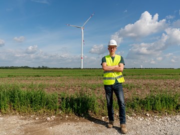 Mitarbeiter vor einer Windkraftanlage Eine Person mit Schutzhelm und gelber Warnweste steht mit verschränkten Armen auf einem Feldweg. Hinter ihr erstreckt sich ein großes grünes Feld mit einem einzelnen Windrad unter blauem Himmel mit Wolken.