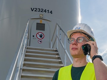 Mitarbeiter im Windpark Eine Person mit Schutzhelm und gelber Warnweste steht vor einem Windkraftanlagen-Turm und telefoniert. Hinter ihr führt eine Metalltreppe zum Eingang des Turms.