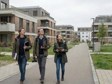 Drei Personen in einem Wohngebiet Drei Personen gehen eine breite Fußgängerallee in einem modernen Wohngebiet entlang. Sie tragen Jacken und halten Unterlagen oder Tablets in den Händen. Rechts und links stehen mehrstöckige Wohngebäude mit Balkonen. Der Himmel ist bedeckt.