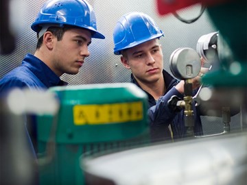 Zwei Personen arbeiten in einem technischen Raum Zwei Personen mit blauen Schutzhelmen und dunkelblauer Arbeitskleidung arbeiten in einem technischen Raum. Sie bedienen Messgeräte und Ventile an einer Maschine. Im Vordergrund sind verschiedene Rohre und Geräte zu sehen.