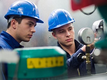 Zwei Personen arbeiten in einem technischen Raum Zwei Personen mit blauen Schutzhelmen und dunkelblauer Arbeitskleidung arbeiten in einem technischen Raum. Sie bedienen Messgeräte und Ventile an einer Maschine. Im Vordergrund sind verschiedene Rohre und Geräte zu sehen.