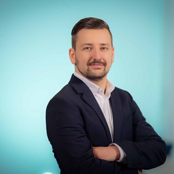 Alexander Rueffler Eine Person steht mit verschränkten Armen in einem modernen Büro. Sie trägt einen dunkelblauen Anzug über einem weißen Hemd.