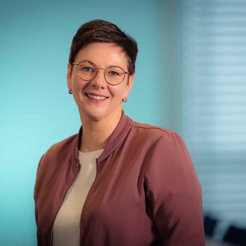 Doreen Graul Eine Person mit kurzen Haaren ist zu sehen. Sie trägt eine weinrote Jacke über einem weißen Oberteil. Der Hintergrund ist unscharf und zeigt einen Farbverlauf von Türkis zu Hellblau.