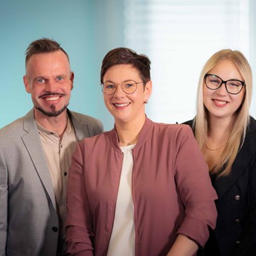 Team Fernwaerme Drei Personen stehen nebeneinander und blicken in die Kamera. Im Hintergrund ist eine türkisfarbene Wand zu sehen. Auf der rechten Seite des Bildes befindet sich ein Fenster, durch das Licht einfällt.