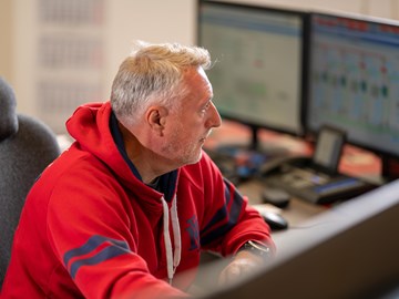 Stoerungsmeldung Eine Person sitzt vor mehreren Computerbildschirmen. Sie trägt einen roten Hoodie mit blauen Akzenten. Der Hintergrund ist leicht unscharf, wodurch der Fokus auf der Person und der Computeranordnung liegt.