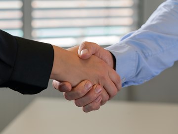 Hände schütteln Nahaufnahme eines Händedrucks zwischen zwei Personen. Eine Person trägt einen schwarzen Anzug, die andere ein hellblaues Hemd. Im Hintergrund sind Fensterjalousien zu sehen, was auf eine Büroumgebung hinweist.